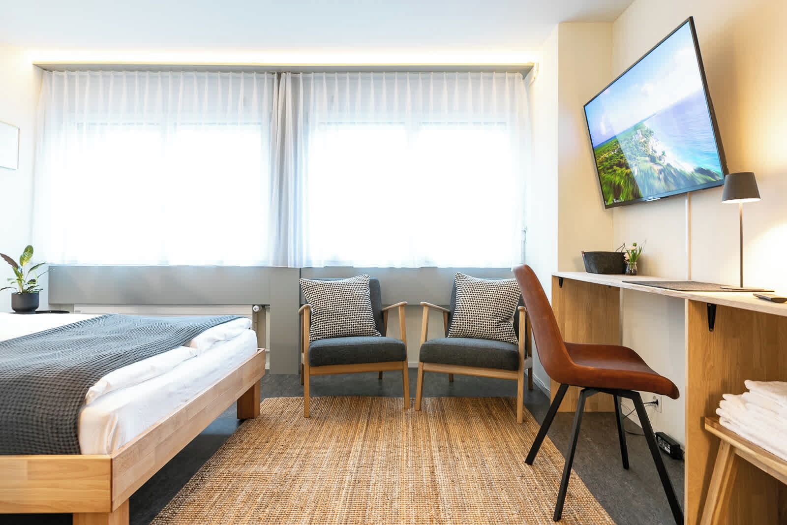 Una habitación de hotel con cama, dos sillas, un escritorio, un televisor y una ventana con cortinas.