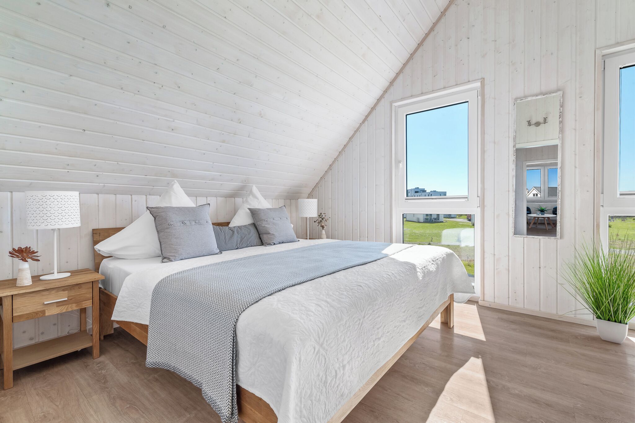A bedroom with a sloped ceiling, a bed with a patterned bedspread, a bedside table with a lamp, and a view of the outdoors through a window.