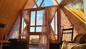 A cozy attic bedroom with wooden walls, a large window with curtains, a wicker chair, and a small table. - Eva Cabins (Dalhousie)