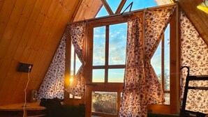 A cozy attic room with wooden walls and a sloped ceiling, featuring a large window with patterned curtains, a wicker chair, and a small table with a lamp. - Eva Cabins (Dalhousie)