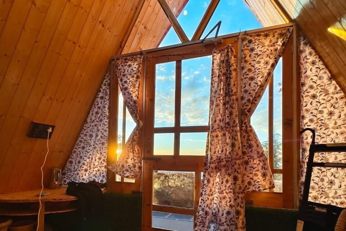 A cozy attic room with wooden walls and a sloped ceiling, featuring a large window with patterned curtains, a wicker chair, and a small table with a lamp.