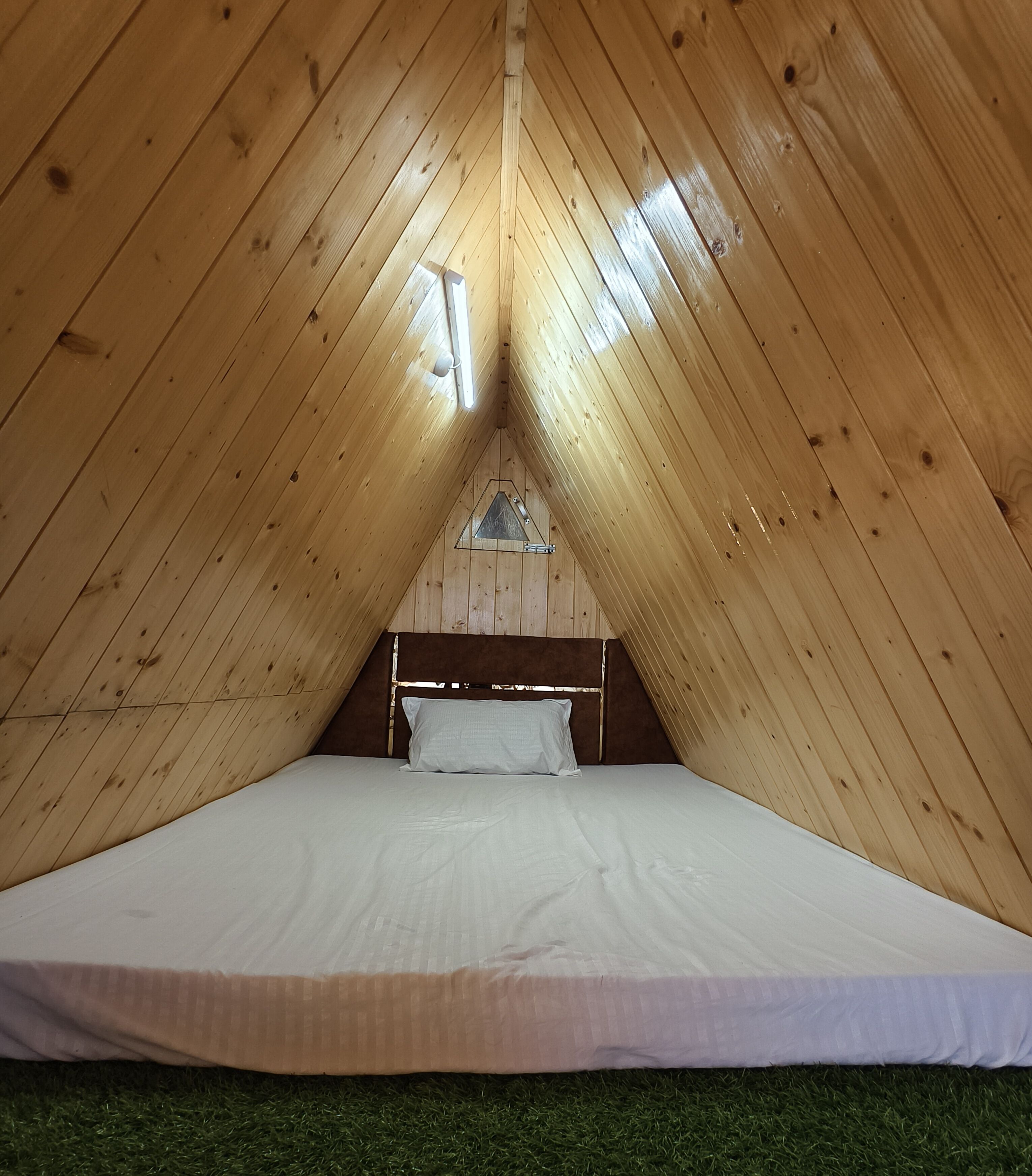 A triangular wooden tent with a bed, a wooden headboard, and a small window.