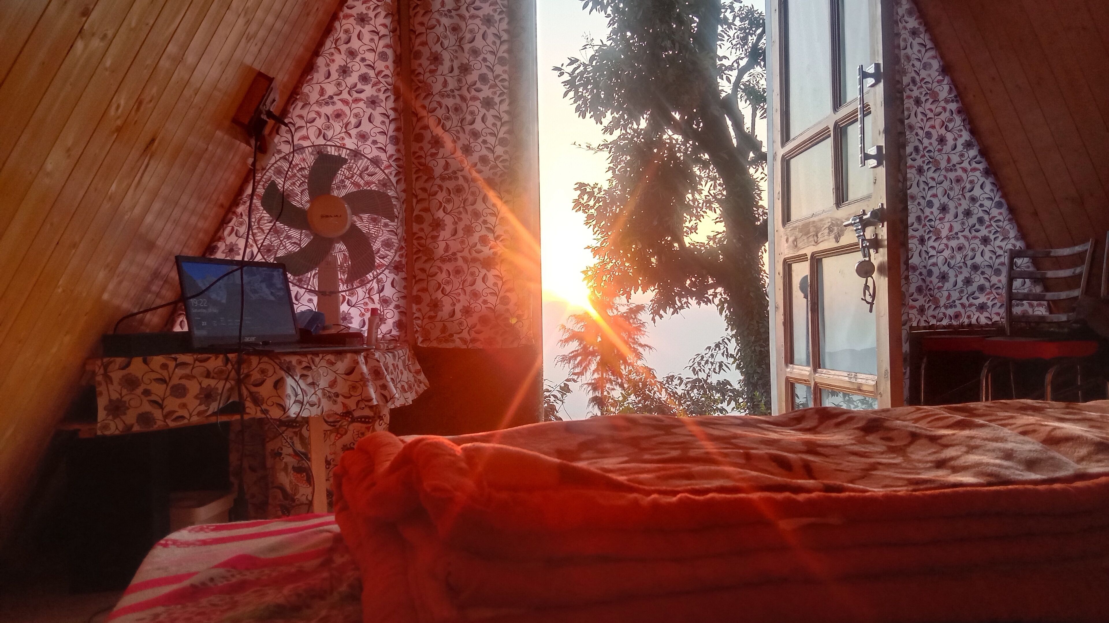 A cozy room with a bed, a desk with a laptop, a fan, and a window with a view of trees and the sun.