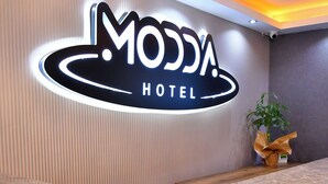 Una recepción con un letrero de neón para Moda Hotel, una planta en maceta y un banco de madera.