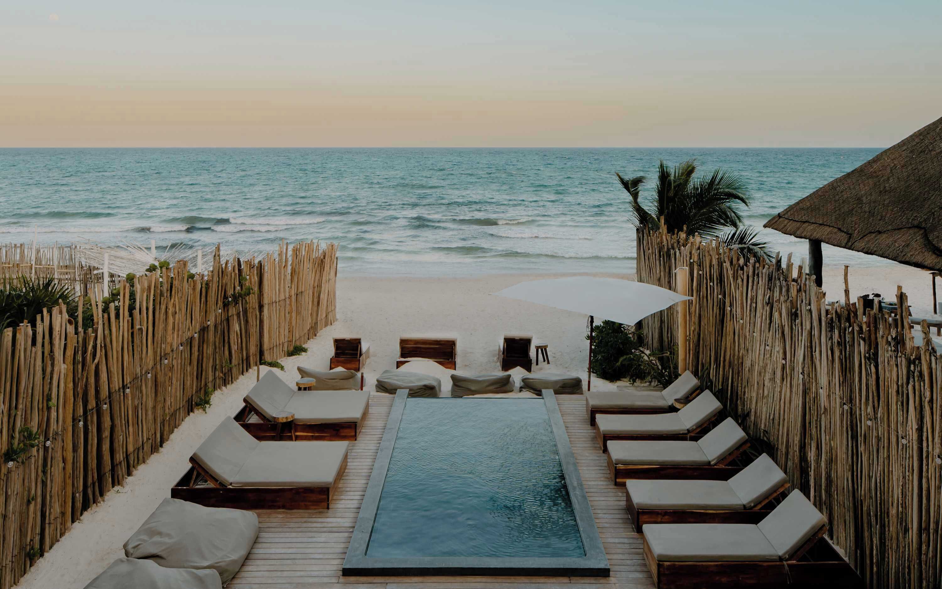 A beachside pool area with lounge chairs and a small pool, surrounded by wooden fences and palm trees.