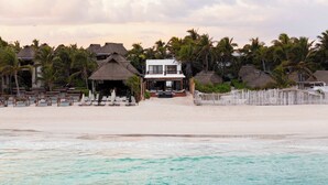 A beachfront property with a large building, multiple thatched-roof structures, and a sandy beach with lounge chairs. - Villa Privada | Fully-Staffed Beachfront Getaway (Tulum)