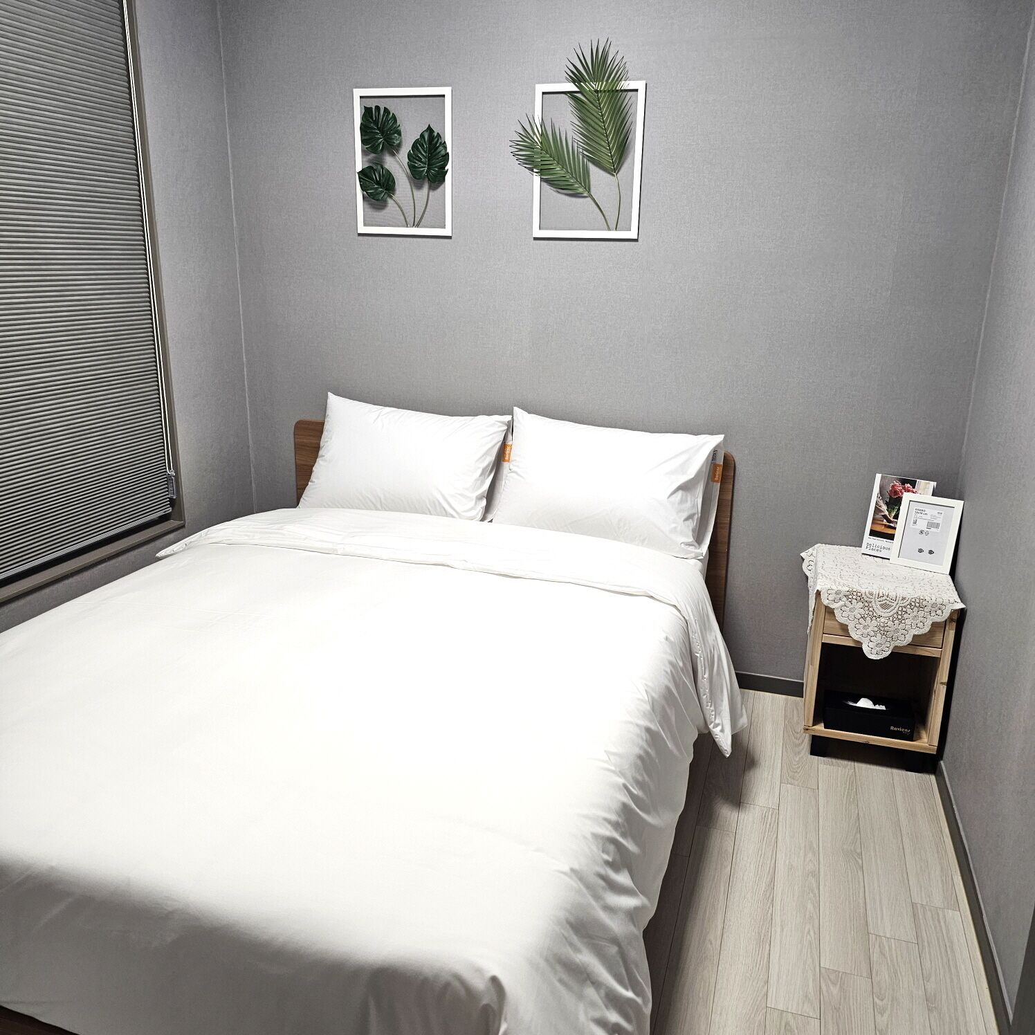 Una cama bien tendida con sábanas blancas en una habitación con paredes grises y suelo de madera. Hay dos cuadros con hojas en la pared sobre la cama.