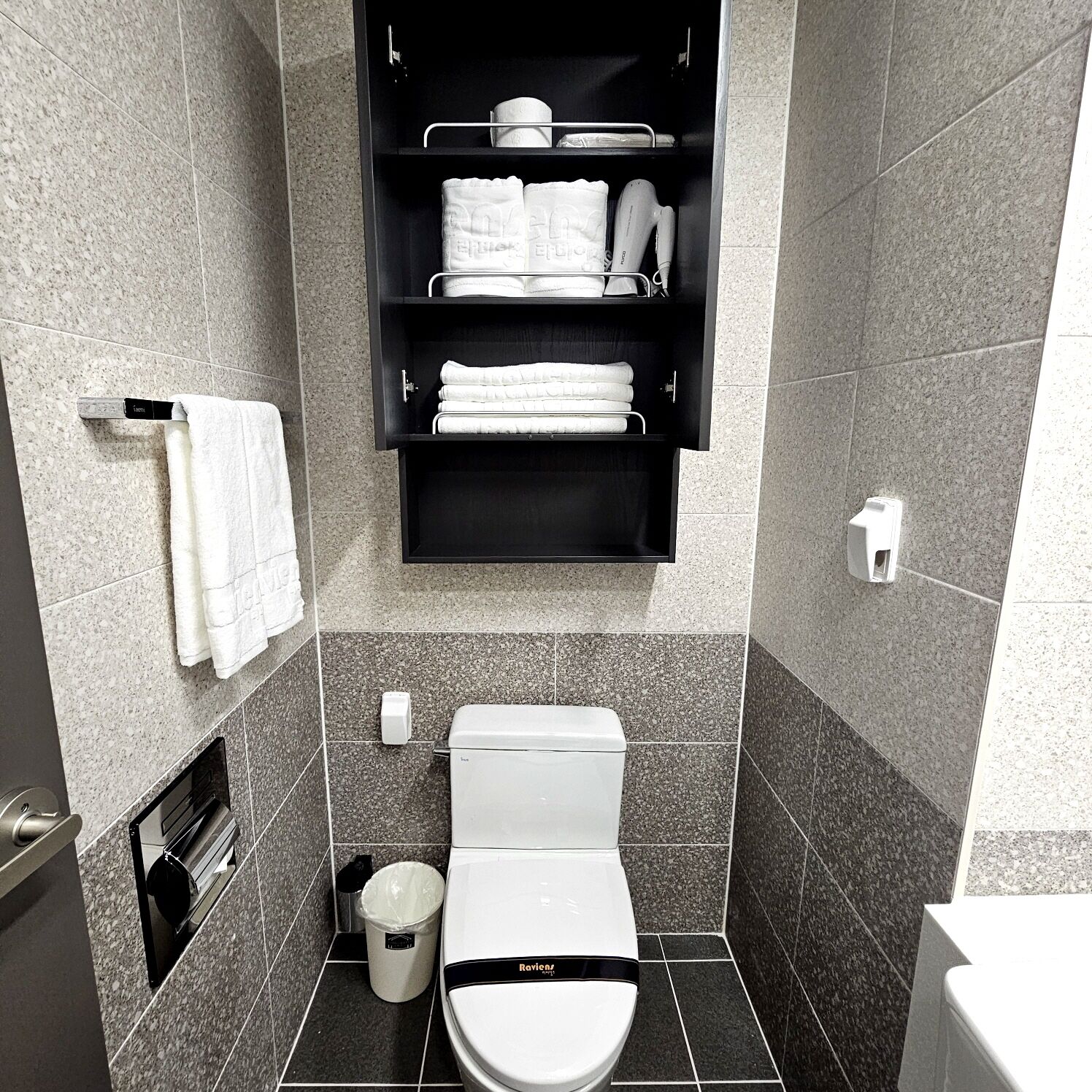A modern bathroom with a toilet, a trash bin, a towel rack, and a shelf with towels and toiletries.