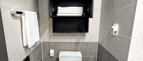 A modern bathroom with a toilet, a trash bin, a towel rack, and a shelf with towels and toiletries.