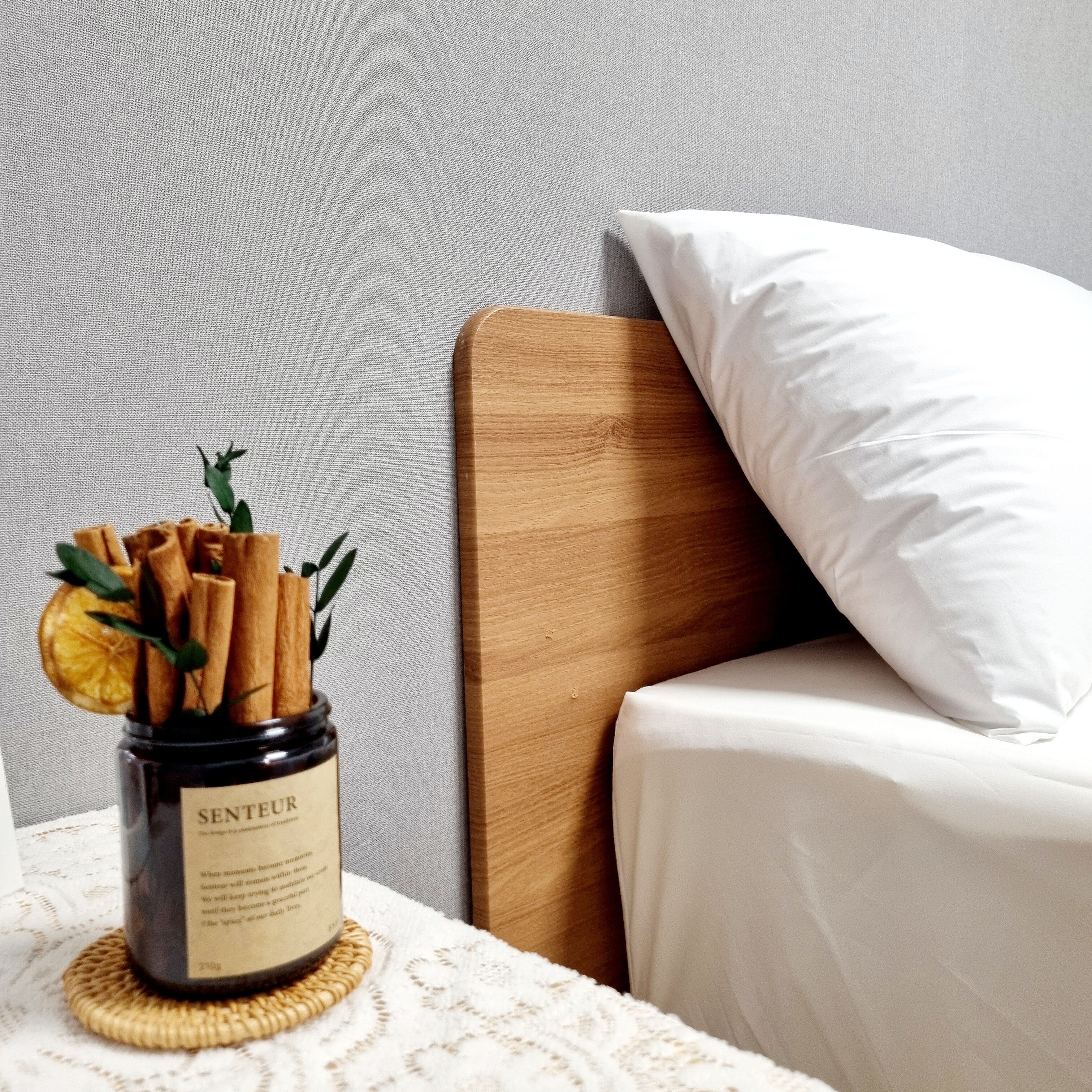 Una mesita de noche con un tarro de ramas de canela y una rodaja de naranja deshidratada, un almohá de color blanco y un cabecero de madera.
