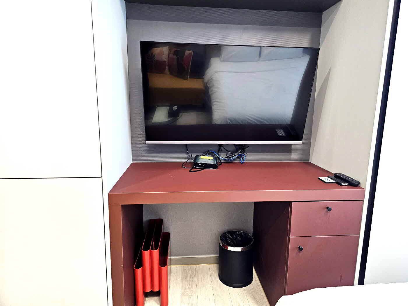 Habitación de hotel compacta con televisión de pantalla plana en la pared, un escritorio pequeño con control remoto y un cesto de basura.