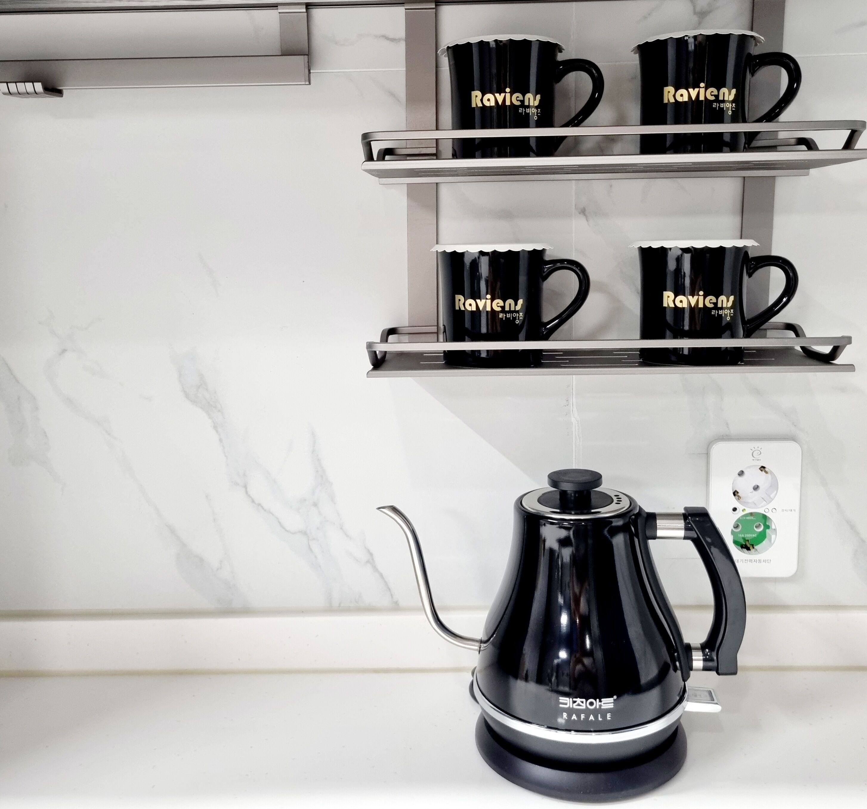 A kitchen with a black coffee maker on the counter, two black mugs on a shelf above, and a white marble backsplash.