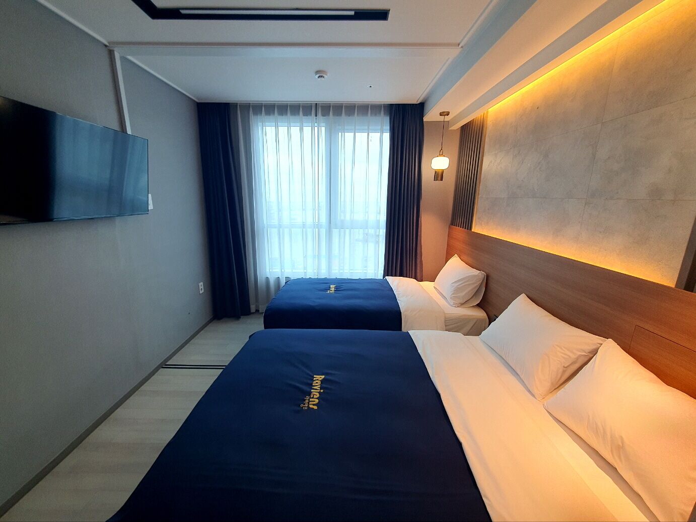 A hotel room with two beds, a flat-screen TV mounted on the wall, and a large window with curtains.