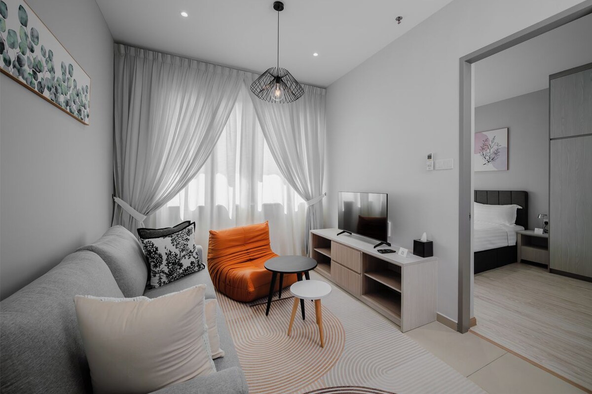 A modern hotel room with a grey sofa, an orange armchair, a small round table, a TV, and a bed with a grey headboard.