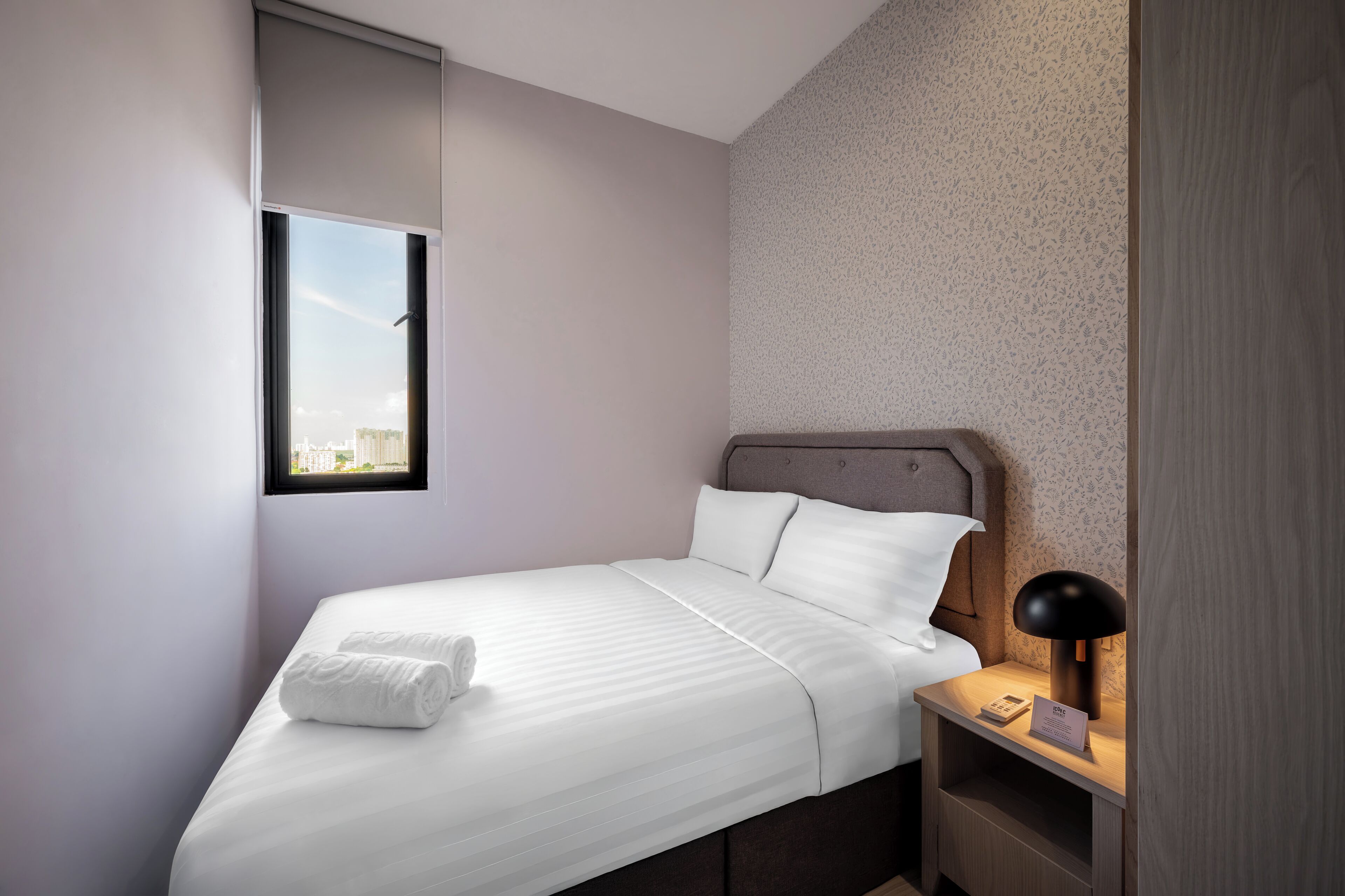 A neatly made bed with white linens and a rolled towel, a bedside table with a lamp and a phone, and a window with a view of buildings.