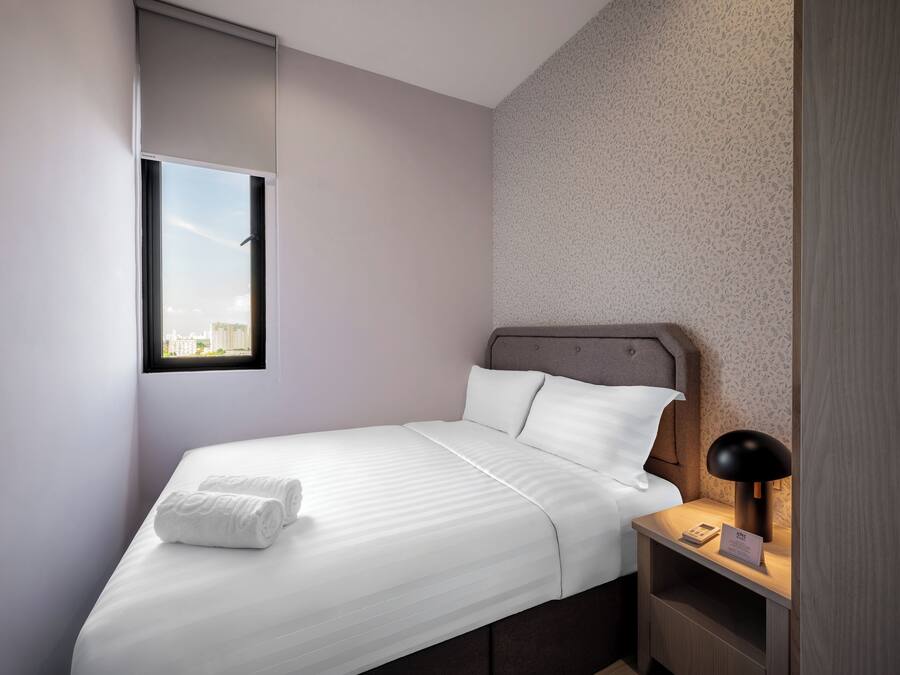 A neatly made bed with white linens and a rolled towel, a bedside table with a lamp and a phone, and a window with a view of buildings.
