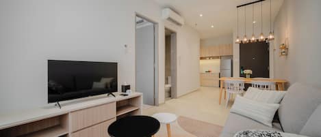 A modern living room with a flat-screen TV, a sofa, a dining area with a table and chairs, and a kitchen in the background.