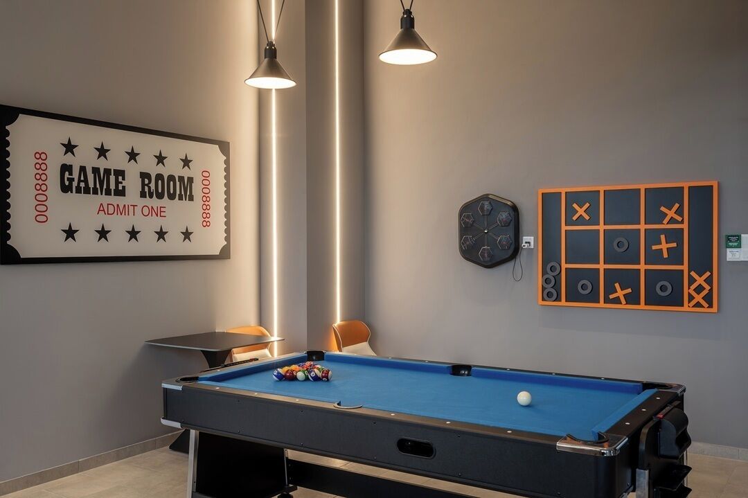 Ein Spielzimmer mit Billardtisch, zwei Pendelleuchten, einer Dartscheibe und einem Wandzeichen mit der Aufschrift 'GAME ROOM ADMIT ONE'.