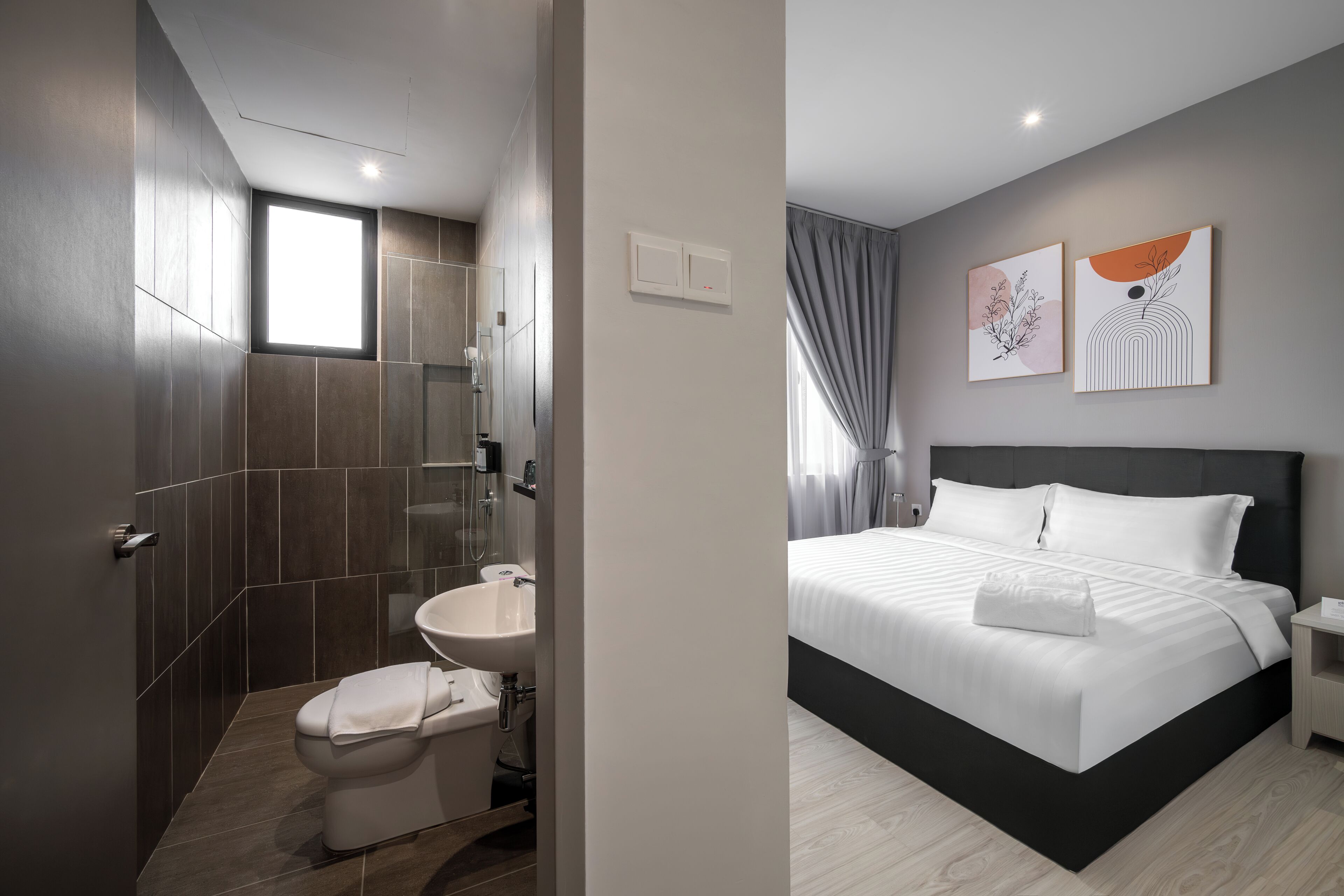 A modern bathroom with a toilet, sink, and shower. The room has a large window and a bed with white bedding.