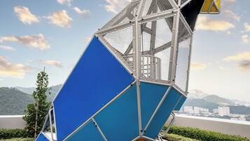 Una escultura de pájaro metálico de diseño geométrico, sobre una base azul y amarilla, ubicada sobre un suelo con estampado azul y verde, con un paisaje urbano al fondo.