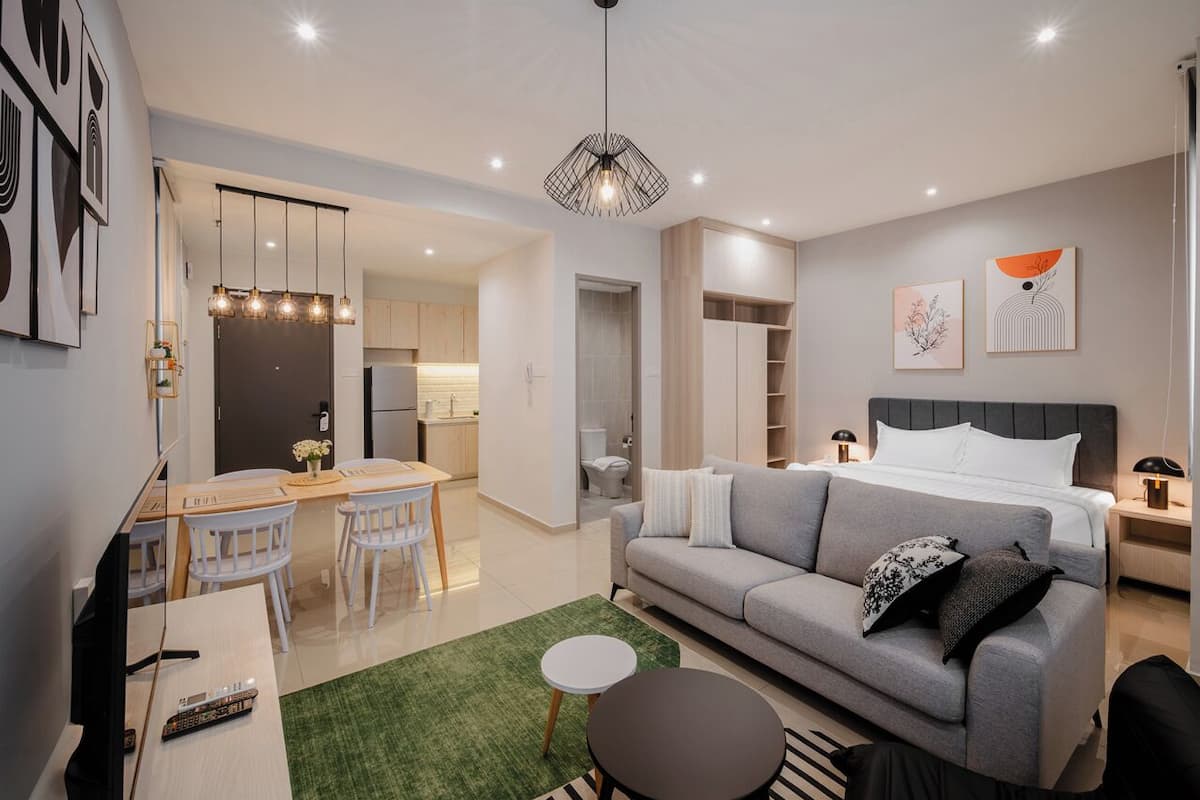 A modern living room with a grey sofa, a dining area with a wooden table and chairs, a kitchen in the background, and a ceiling light fixture.