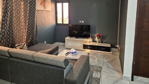 A living room with a grey sofa, a TV on a white stand, a patterned curtain, and a window with natural light. - COSY apartment in Abidjan (Abidjan)