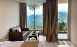 A hotel room with a bed, a sofa, a small table, and a view of mountains through large windows.