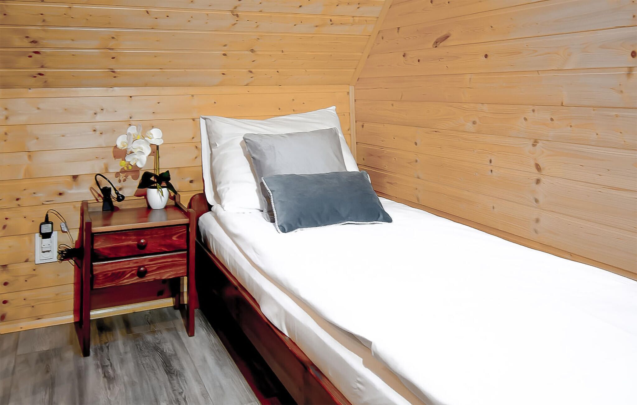 A wooden cabin bedroom with a bed, a nightstand, a lamp, and a vase with flowers.