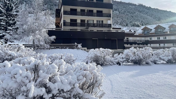 Ein modernes, mehrstöckiges Gebäude mit Balkonen, umgeben von schneebedeckten Bäumen und einer Bergkulisse.