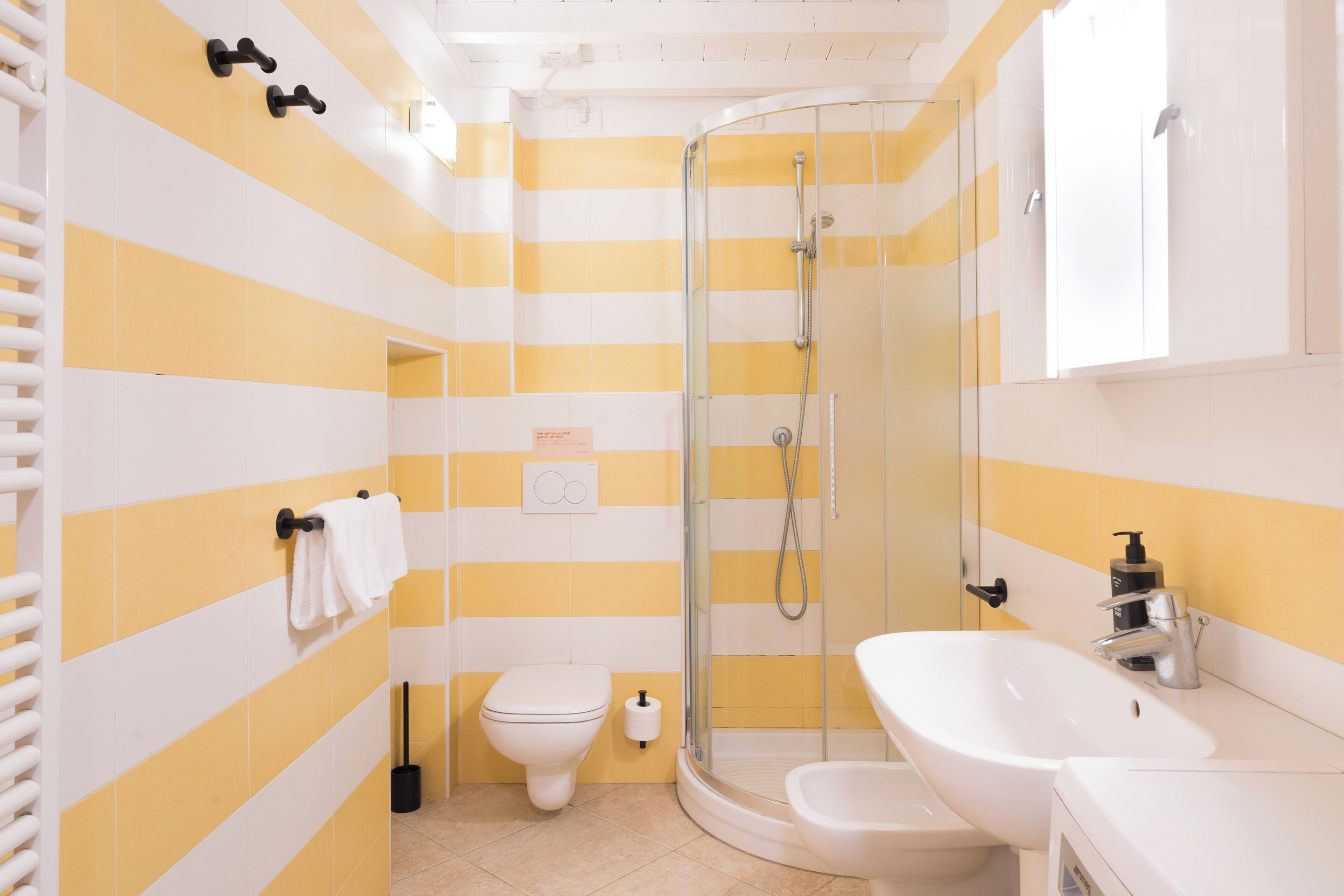 Un baño con paredes a rayas amarillas y blancas, un box de ducha de vidrio, un inodoro blanco y un lavabo blanco con un espejo encima.