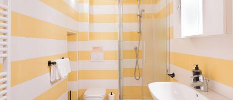 Un baño con paredes a rayas amarillas y blancas, un box de ducha de vidrio, un inodoro blanco y un lavabo blanco con un espejo encima.