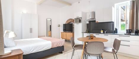 Een moderne hotelkamer met een bed, nachtkastje, lamp en een kitchenette met een kookplaat, magnetron en kastjes.