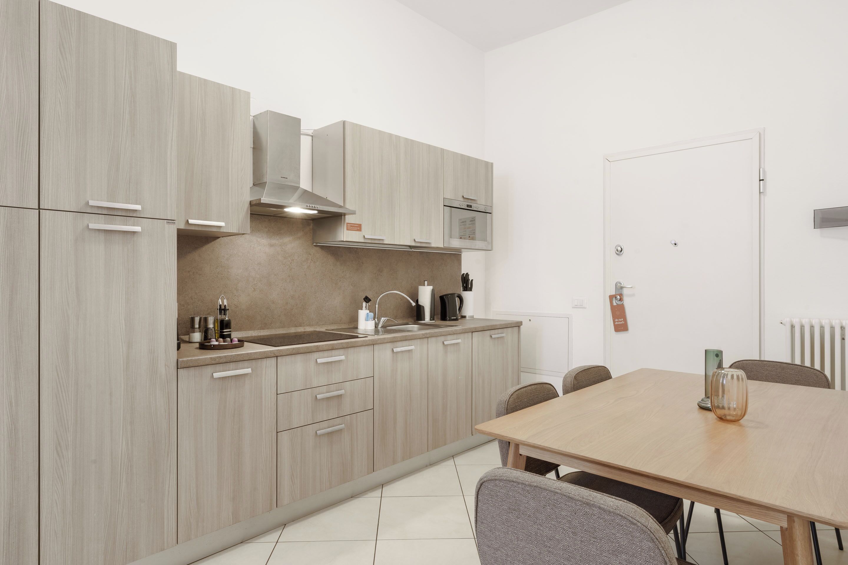 A modern kitchen with light wood cabinetry, a stainless steel range hood, a microwave, and a dining area with a wooden table and chairs.