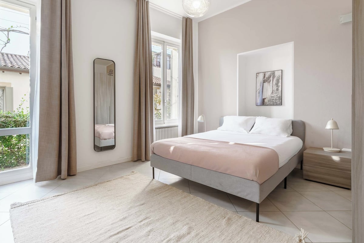 Ein Schlafzimmer mit Bett, Nachttisch, Lampe, Spiegel und einem Fenster mit Vorhängen.