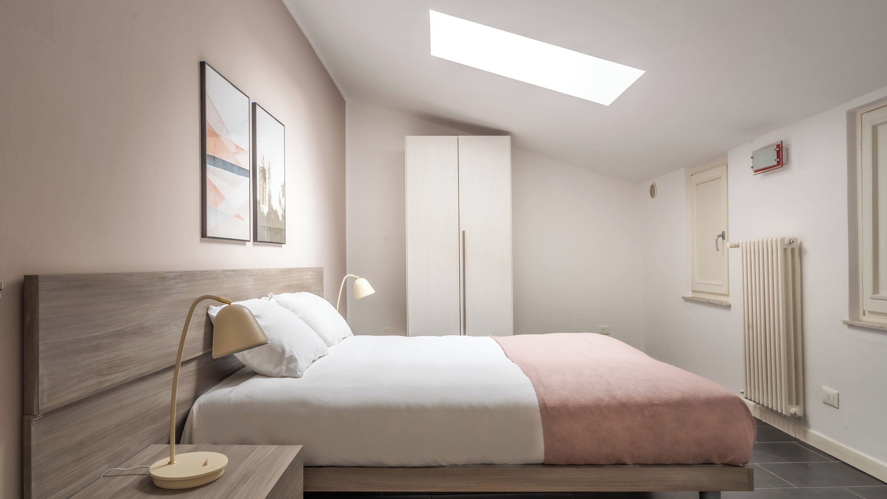 A bedroom with a bed, bedside table, lamp, wardrobe, radiator, and a skylight.