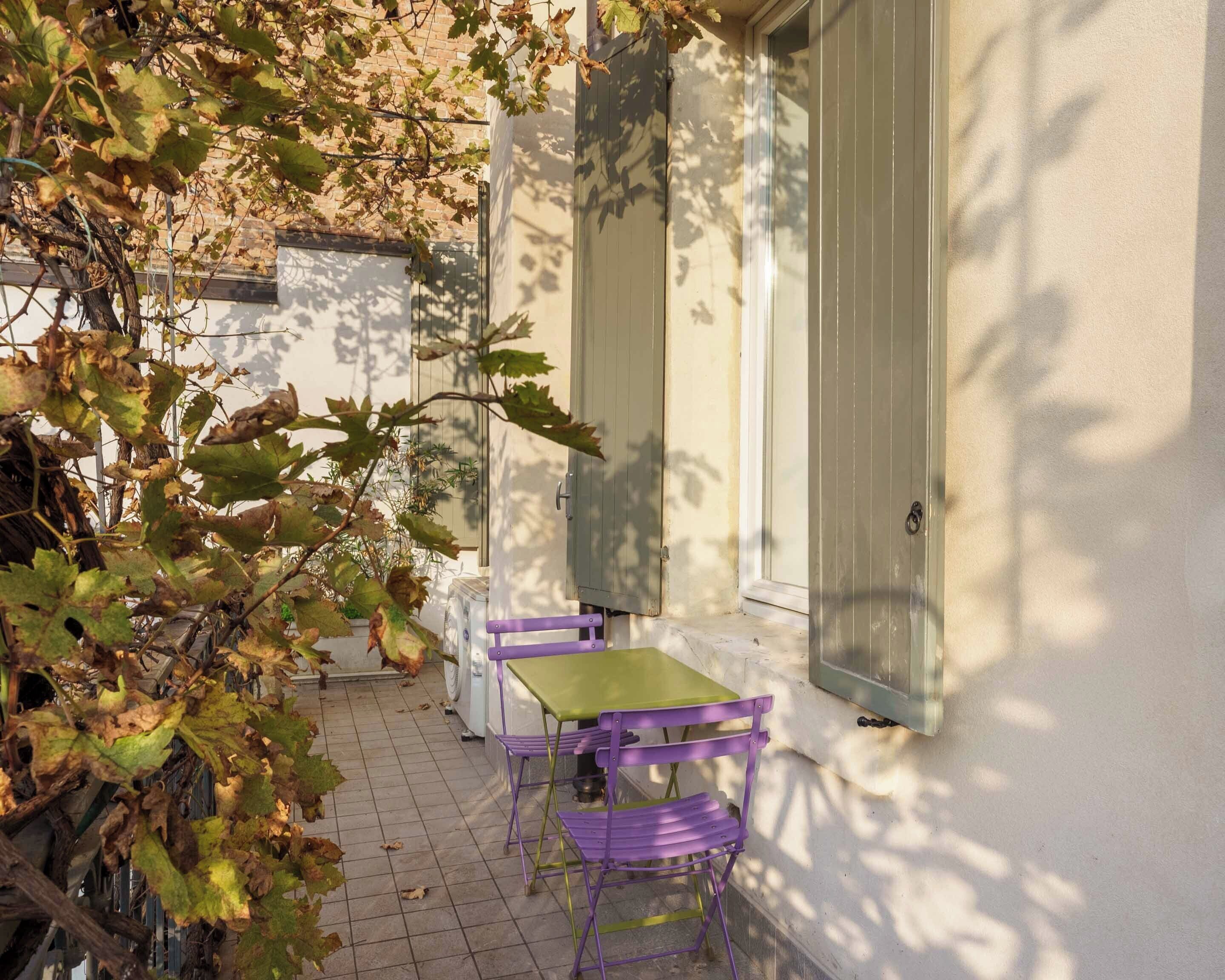 Eine kleine Terrasse mit Tisch und Stühlen, ein Fenster mit Rollläden und eine mit Efeu bewachsene Mauer.