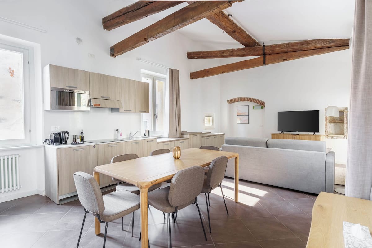 Eine moderne Küche mit Holzbalken, einem Essbereich mit Tisch und Stühlen, einem grauen Sofa und einem Flachbildfernseher.