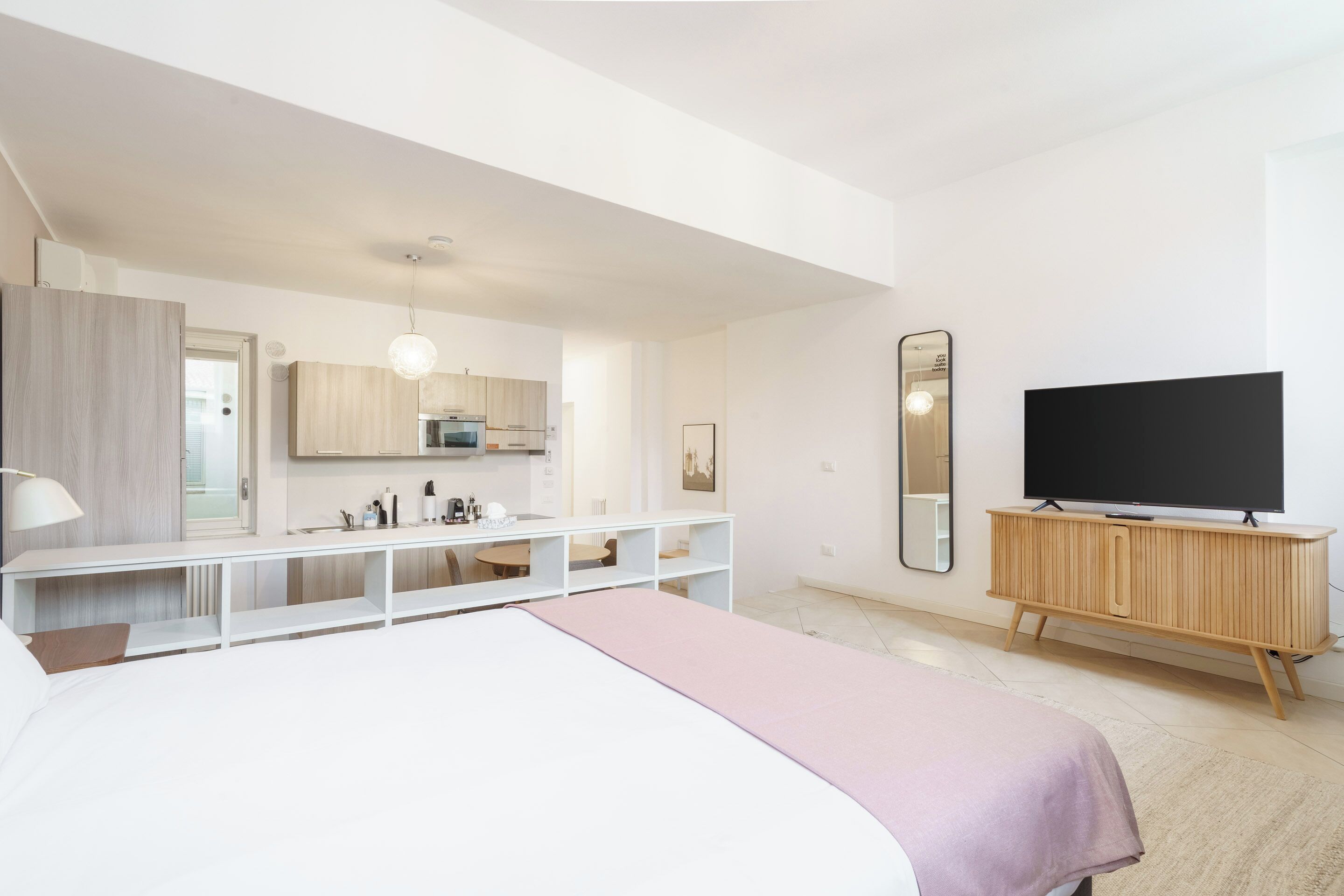 A modern hotel room with a bed, a TV, a wooden cabinet, and a kitchenette.