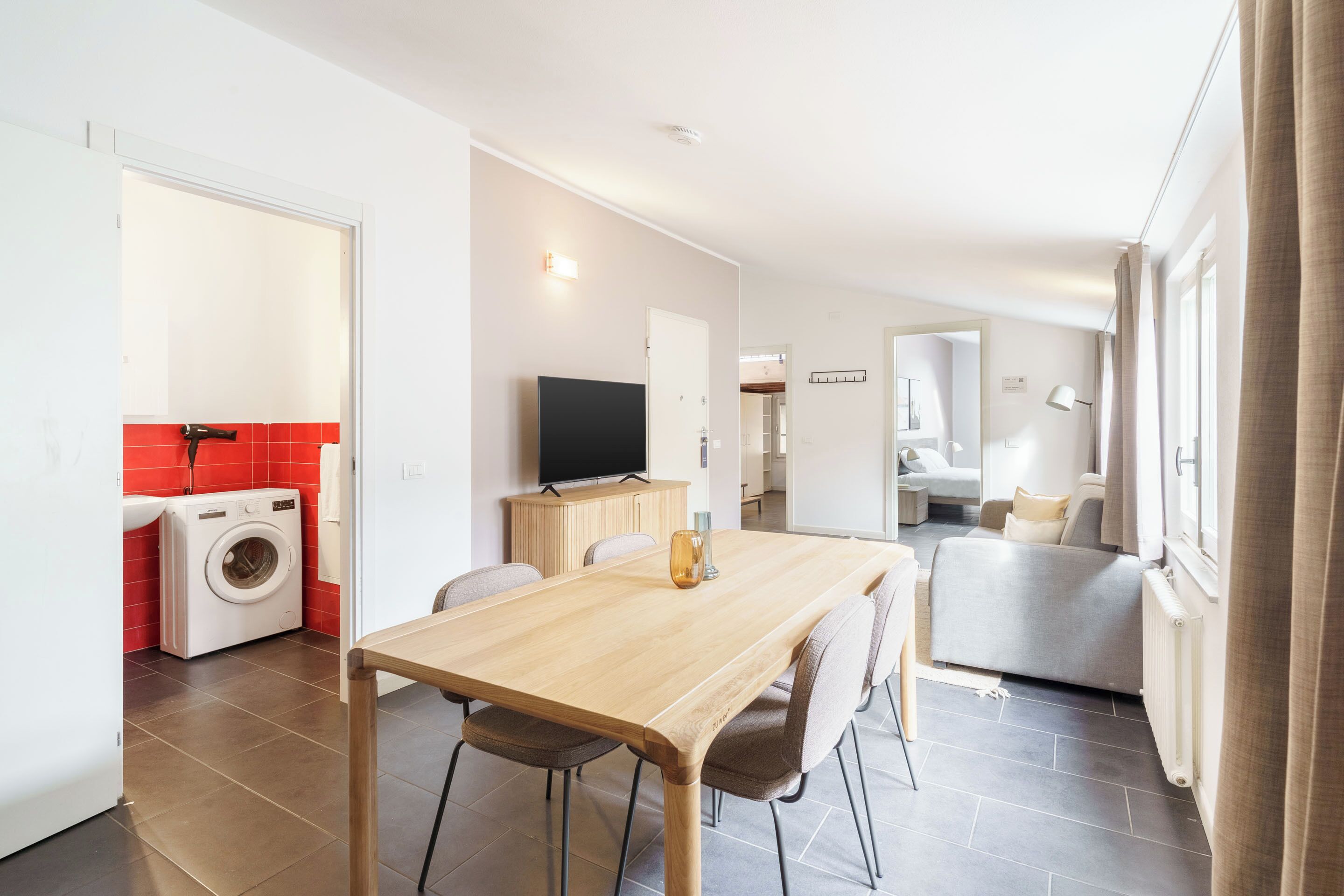 A modern kitchen with a built-in washing machine, a dining area with a wooden table, and a living space with a sofa and a TV.