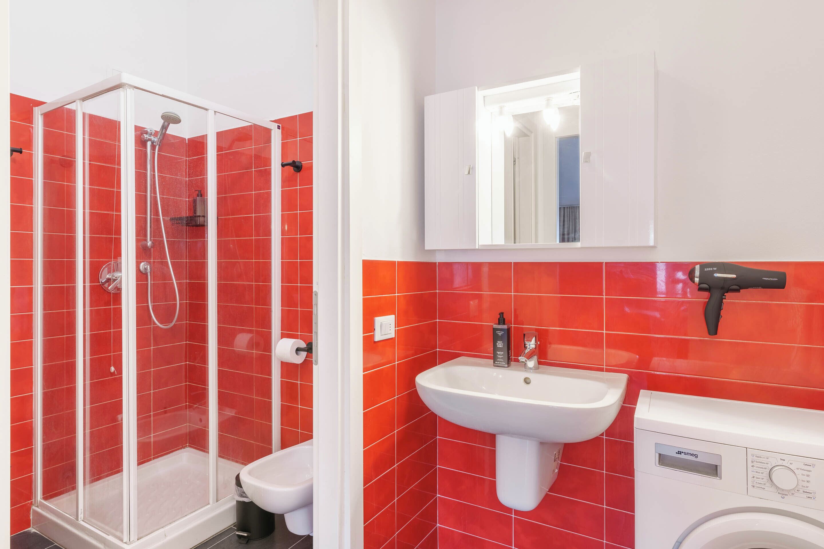 A bathroom with red tiles, a glass-enclosed shower, a sink, a mirror, and a washing machine.