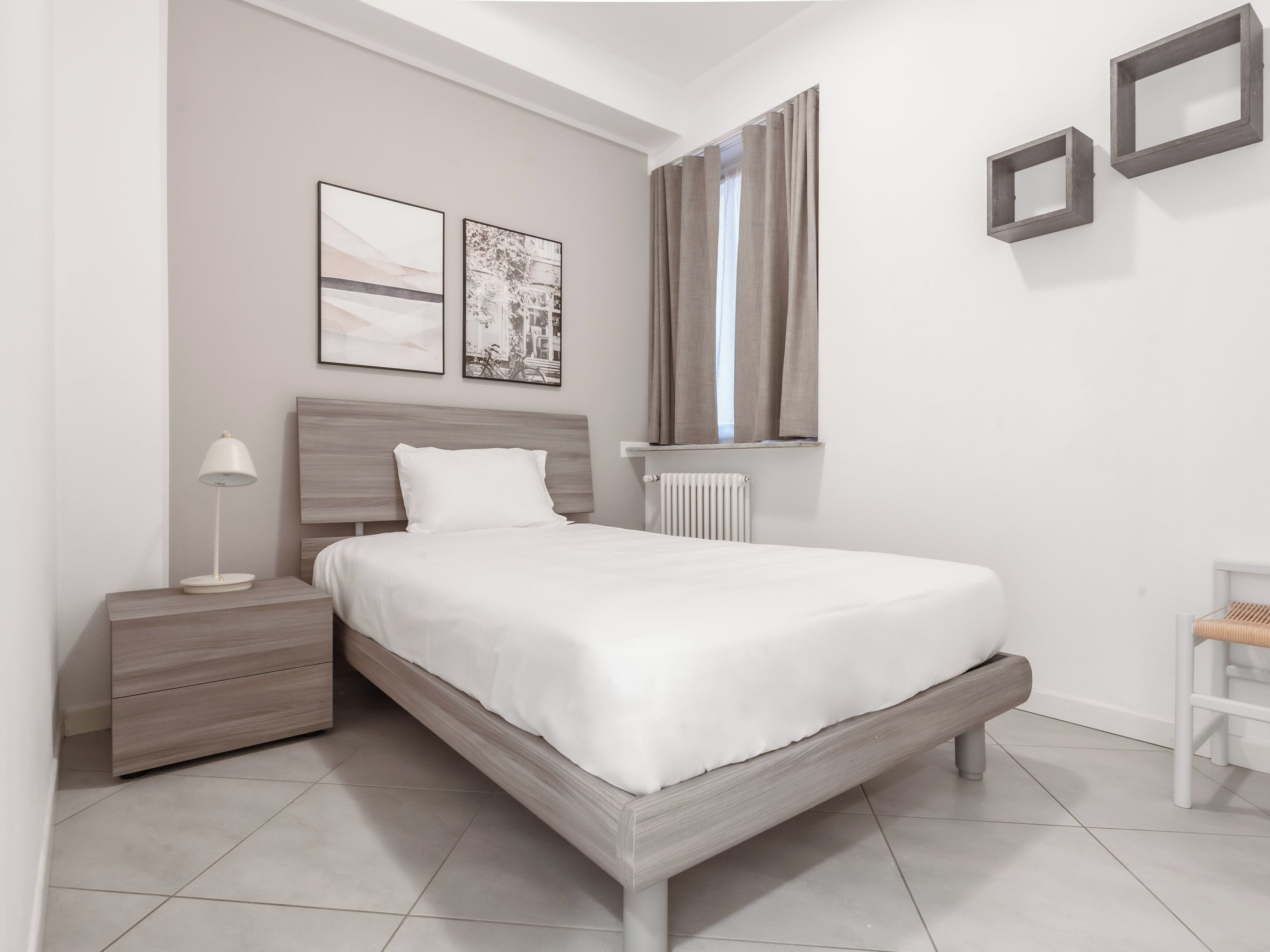 A modern bedroom with a wooden bed frame, a bedside table with a lamp, a radiator, and two framed pictures on the wall.