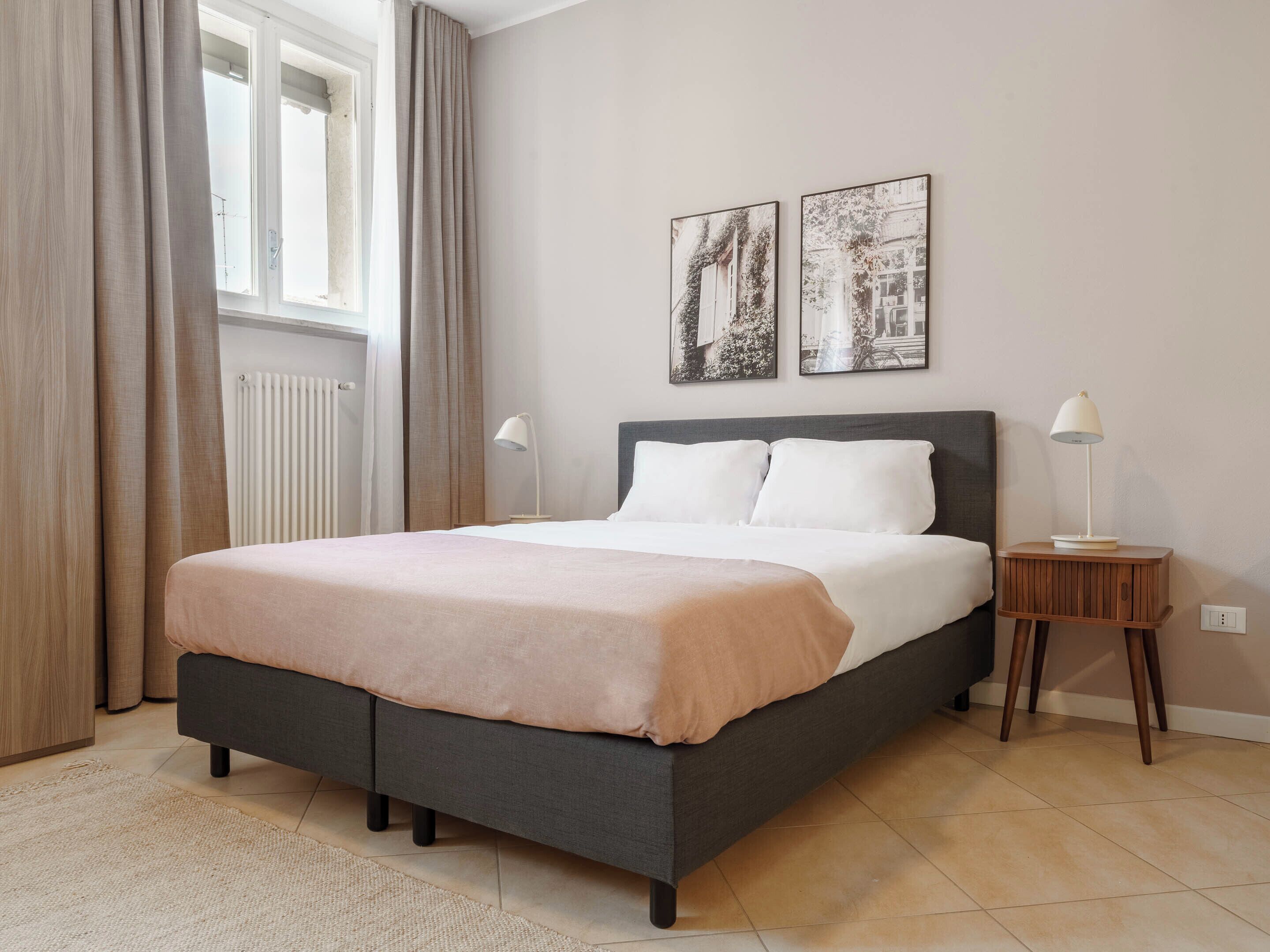 A bedroom with a bed, two bedside tables, a window with curtains, and two wall-mounted pictures.
