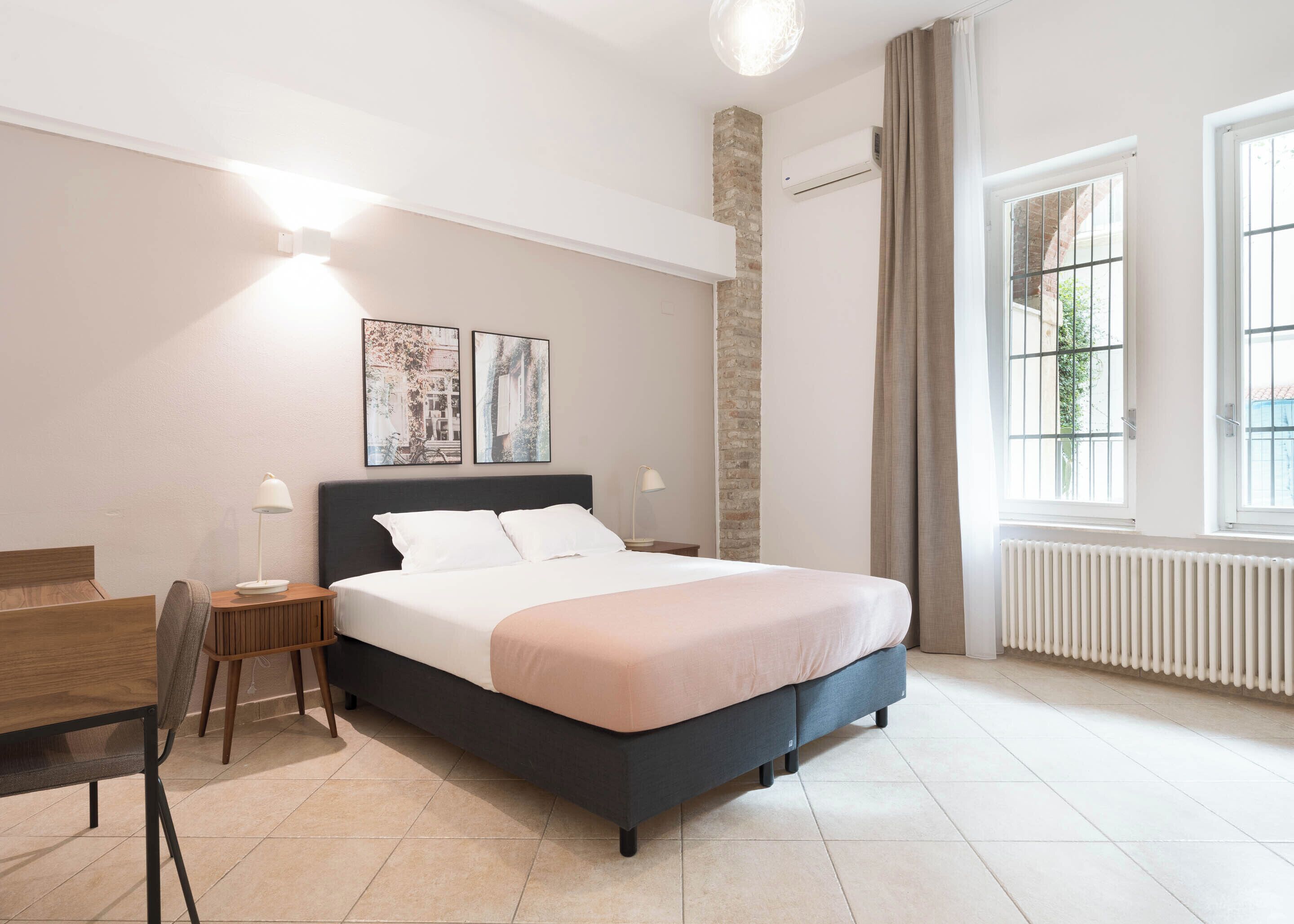 Una camera da letto moderna con un letto, comodini, una scrivania e una sedia. Sono presenti due condizionatori fissati al muro e una finestra con tende.