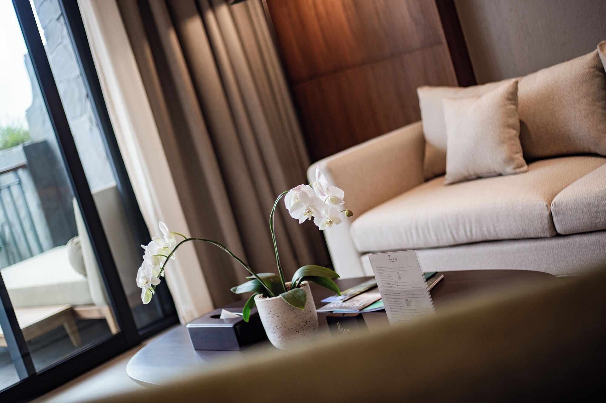 A modern living room with a beige sofa, a small table with a potted orchid, and a window with curtains.