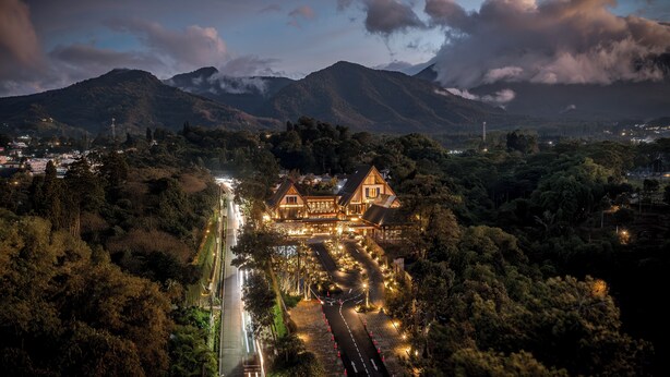 Un grand hôtel illuminé, avec un bâtiment central imposant, entouré d’une végétation luxuriante et dominé par des montagnes en arrière-plan.