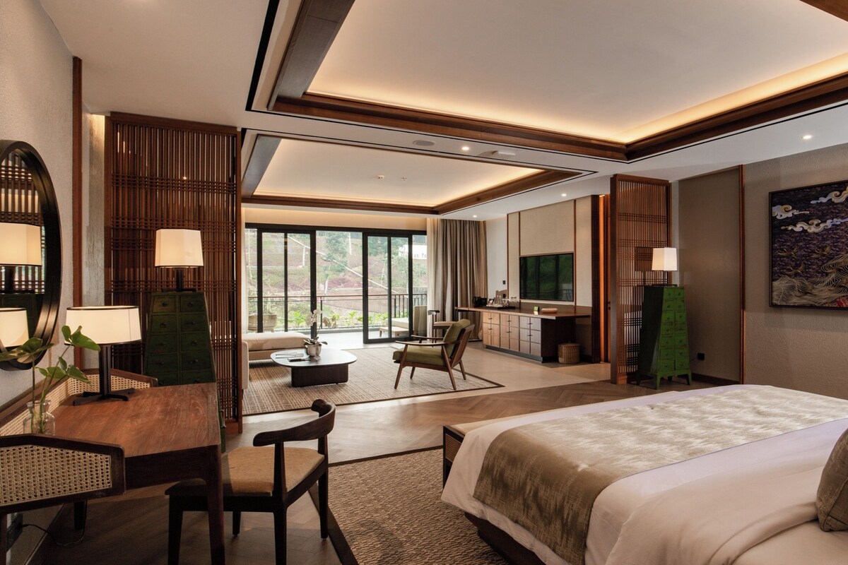 A modern hotel room with a large bed, a desk with a chair, a dining area, and a view of the outdoors.