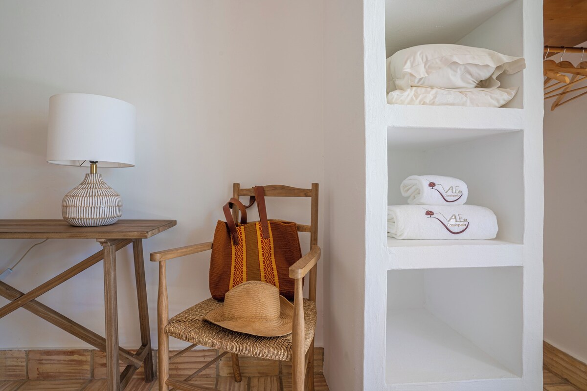 A wooden chair with a woven seat and a straw hat, a white lamp, and a shelf with folded towels and a bag.