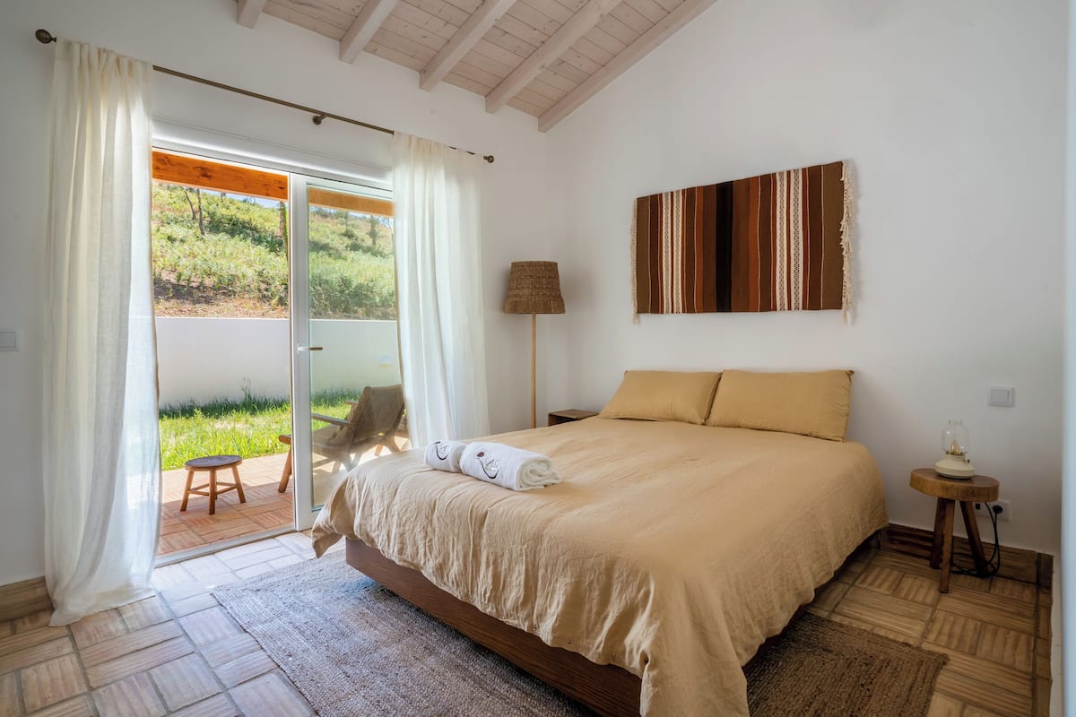 A bedroom with a bed, a nightstand, a lamp, and a view of the outdoors.