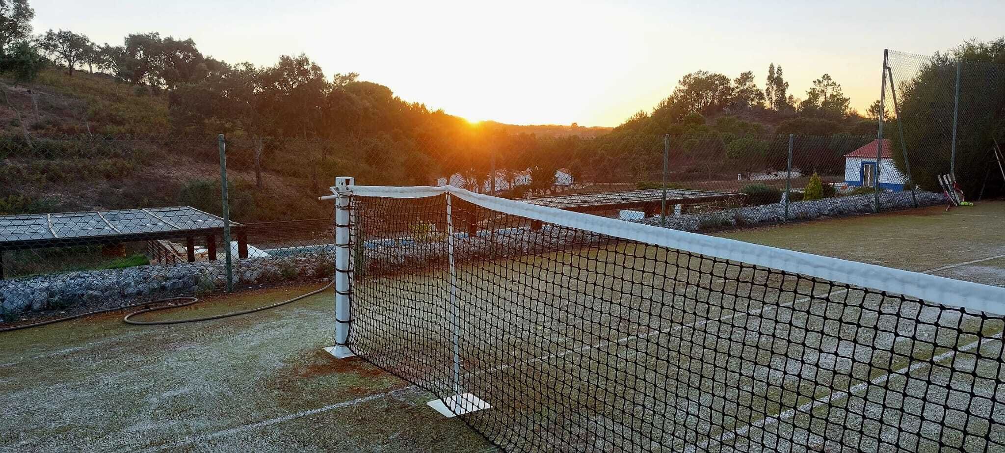 Ein Tennisplatz mit Netz, umzäunt, mit einem kleinen Gebäude im Hintergrund.