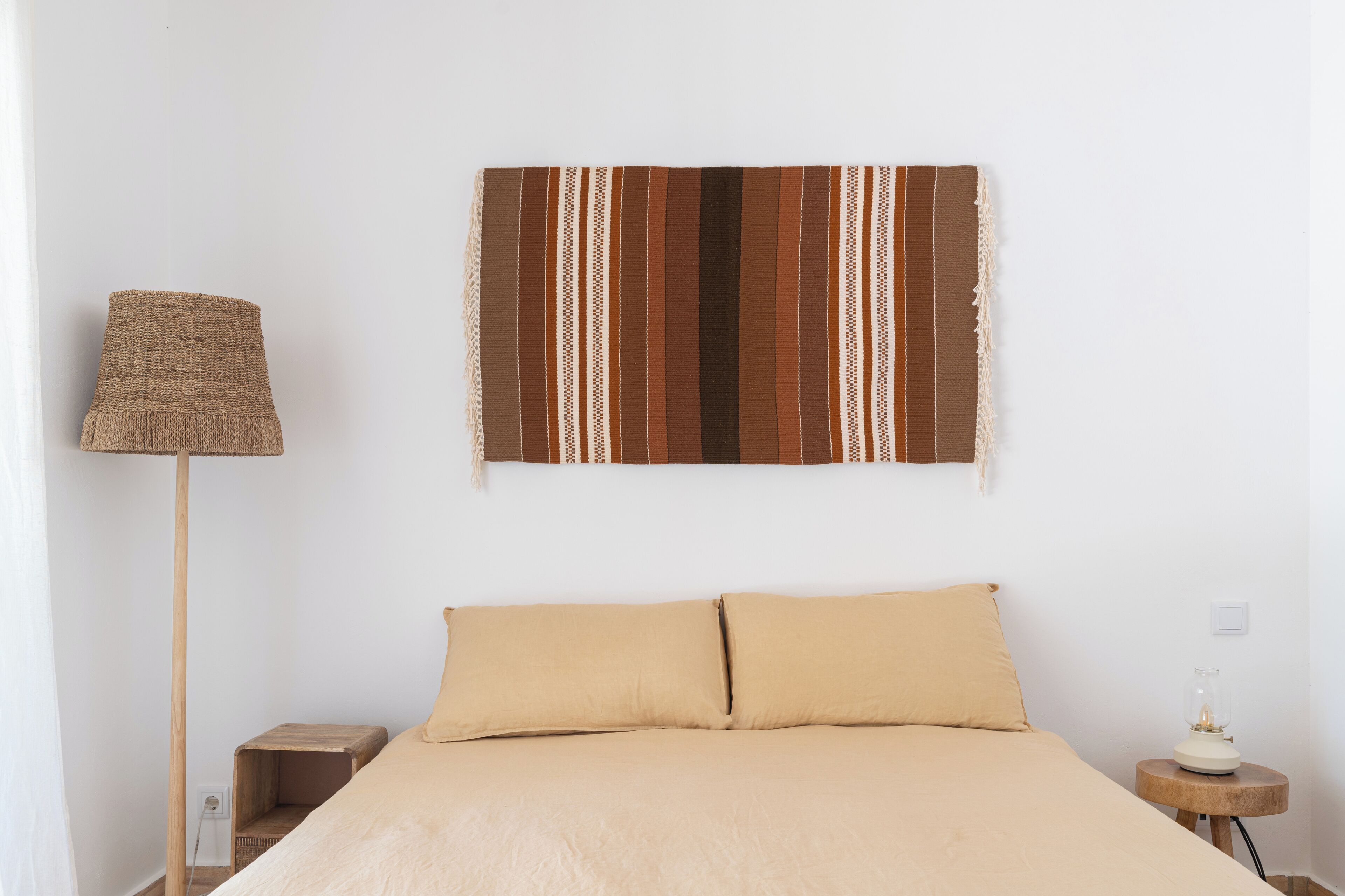 A bedroom with a bed, a wall-mounted striped tapestry, a floor lamp, and a bedside table with a lamp.